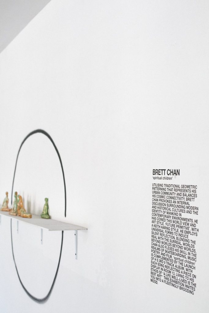 Brett Chan 'Spiritual Children' wall text beside a painted circle and shelf of small figurines at The Slow