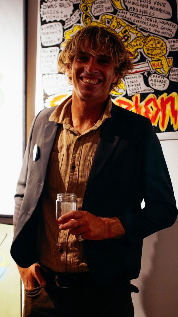 Smiling guest holding a glass in front of illustrated artwork at the Ozzy Wrong opening exhibition, The Slow